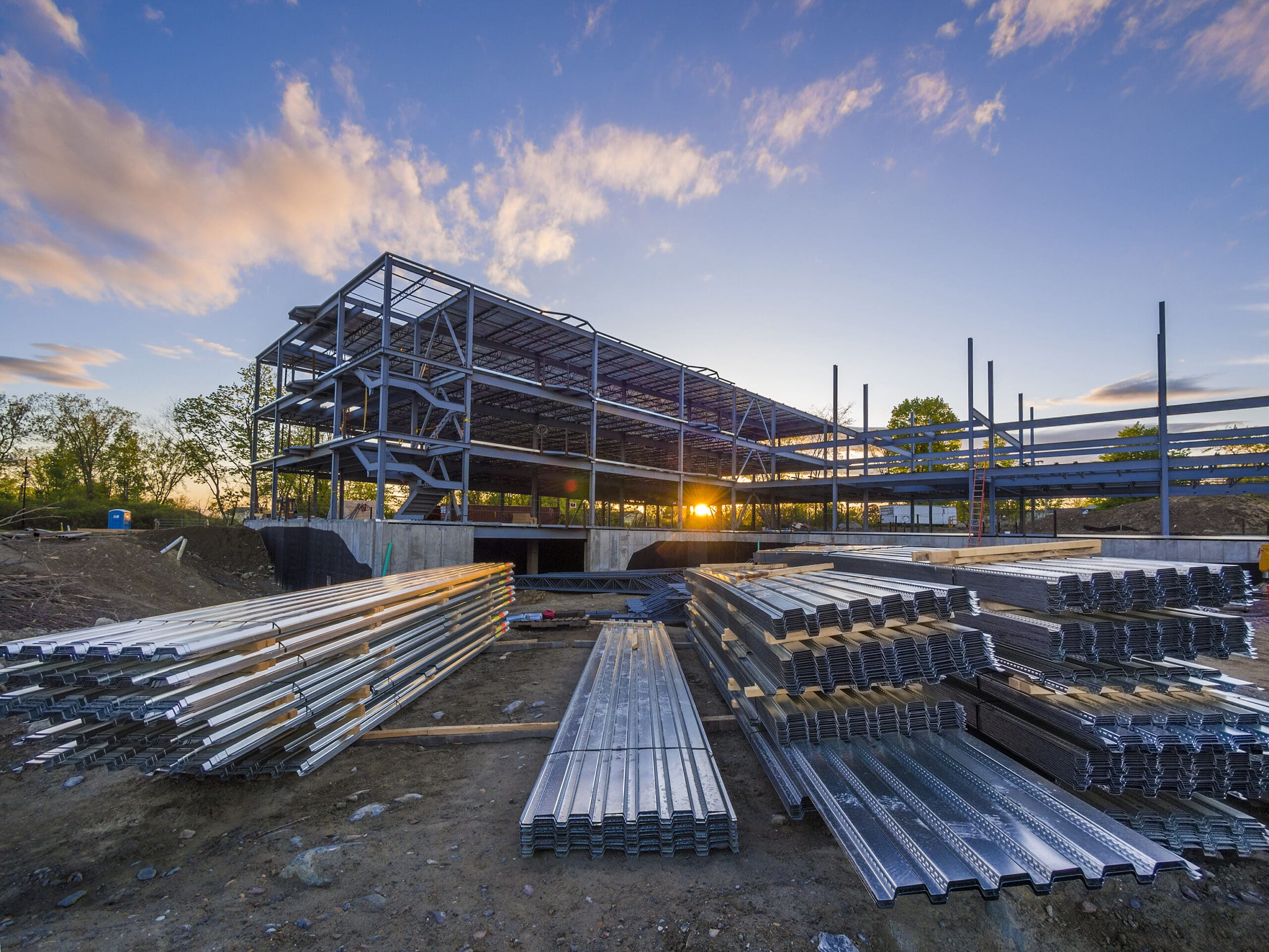 Chantier de construction avec plancher en acier devant un bâtiment partiellement érigé au coucher du soleil.