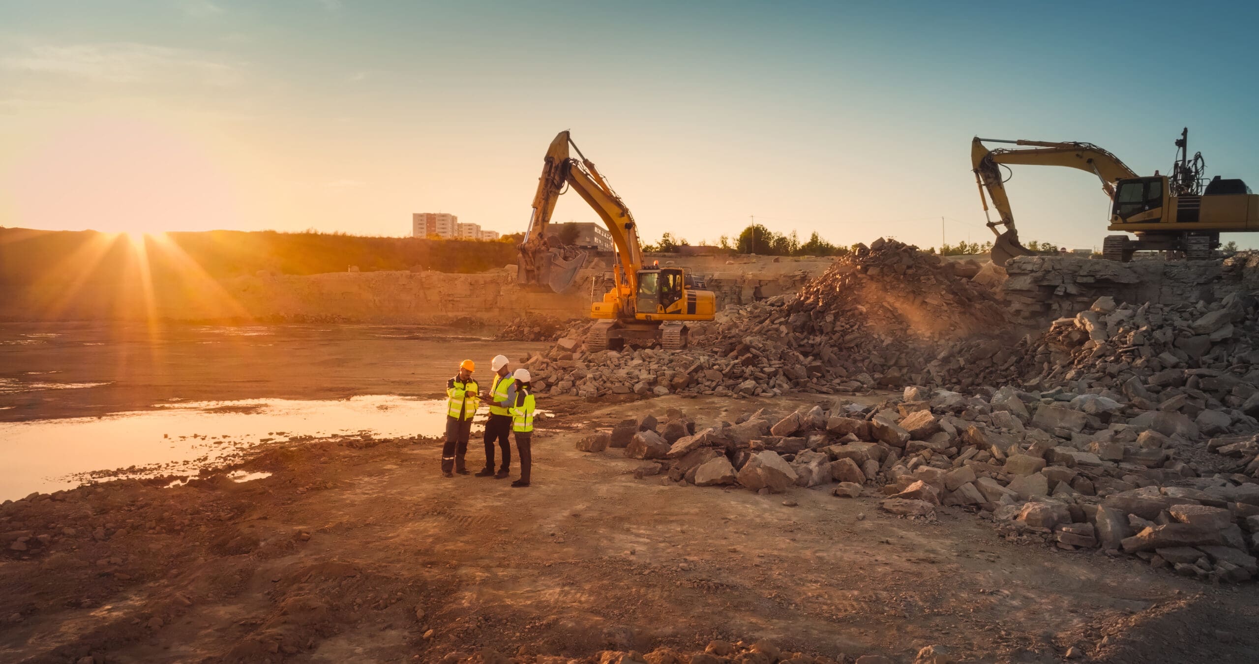 Prise de vue aérienne par drone d’un chantier de construction avec des excavatrices par une journée ensoleillée : une équipe diversifiée de promoteurs immobiliers discute du projet. Un ingénieur civil, un architecte et un inspecteur échangent en utilisant une tablette.