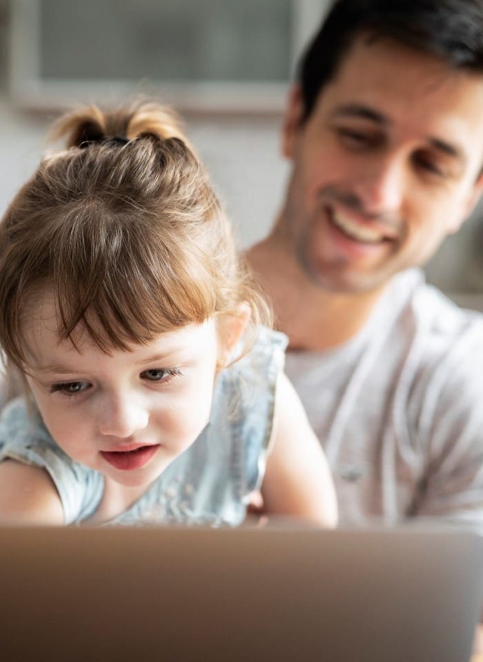 Un père de famille souriant en arrière-plan tenant sa petite fillette dans ses bras devant un ordinateur de travail